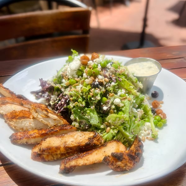 Grilled chicken and a fresh salad with peas, feta, and nuts, served with dressing on a white plate.