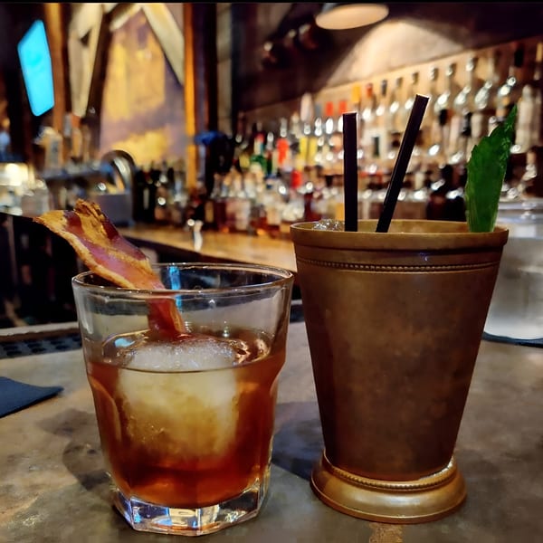 Two cocktails on a bar, one with a bacon garnish, the other with mint.
