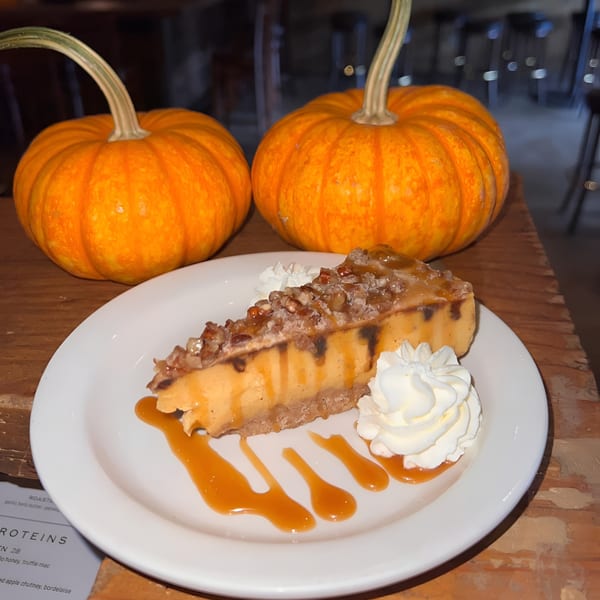 A slice of pumpkin cheesecake with whipped cream and caramel sauce, flanked by two small pumpkins.