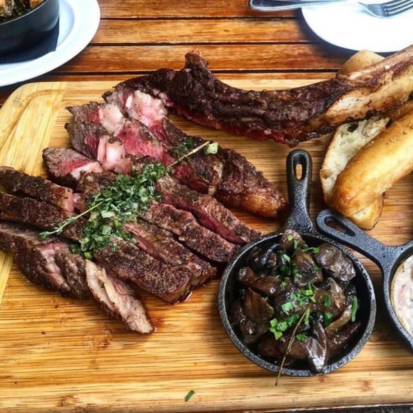 Sliced steak with herbs, mushrooms in cast iron pans, and bread on a wooden board.