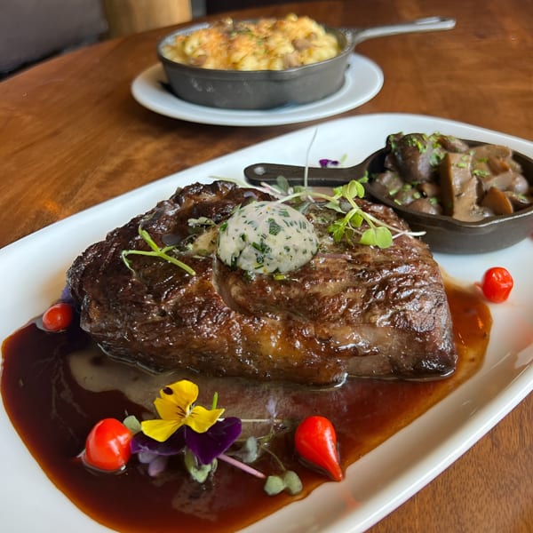 Ribeye steak with herb butter, mushrooms, and mac and cheese on a wooden table.