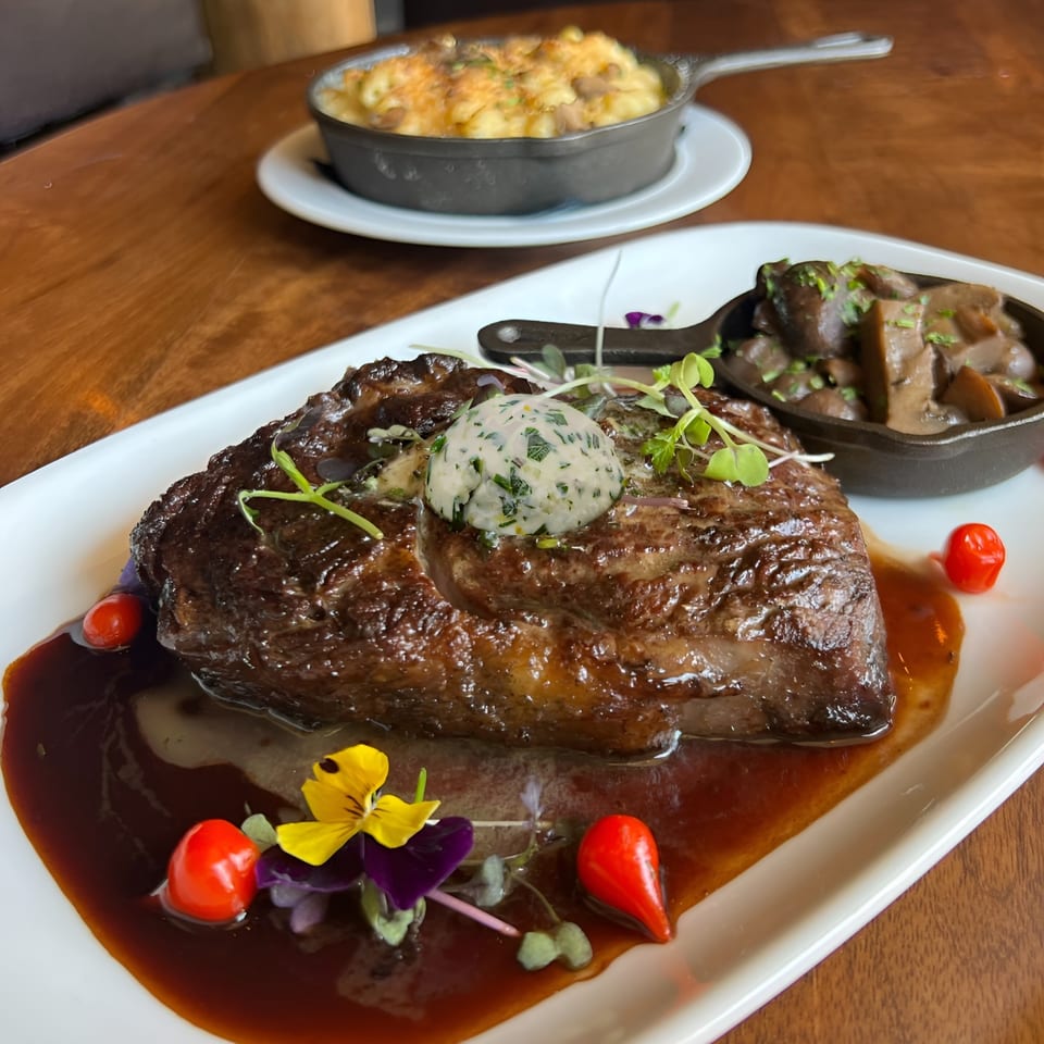 Ribeye steak with herb butter, mushrooms, and mac and cheese on a wooden table.