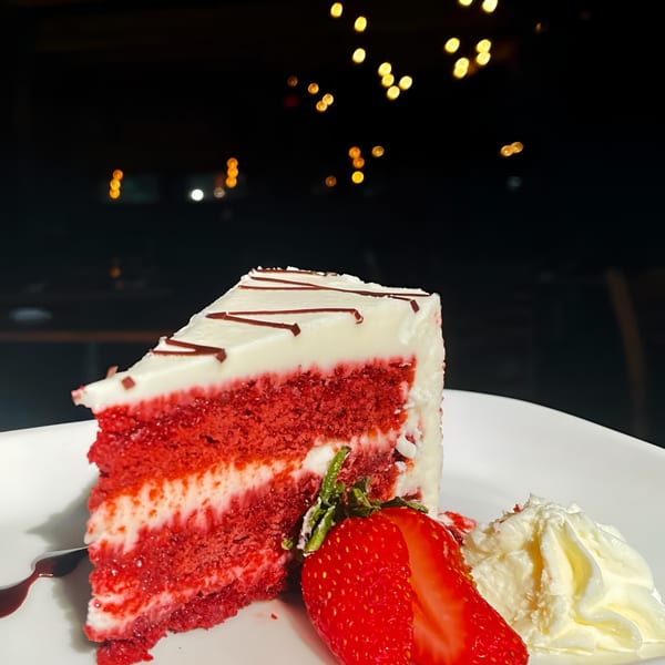A slice of red velvet cake with cream cheese frosting, chocolate drizzle, strawberry, and whipped cream.