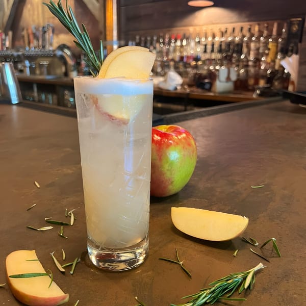 A cocktail garnished with rosemary and pear, with apples and rosemary sprigs on a bar.